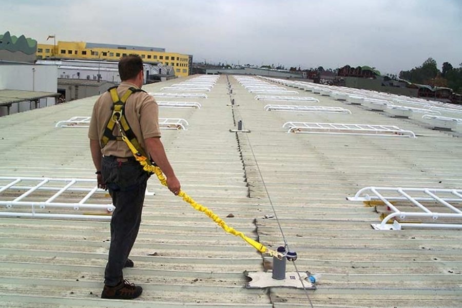 A worker in a fall-arrest harness on a metal industrial roof, connected by a yellow lanyard to a horizontal lifeline anchor system of the kind BSE designs and inspects.